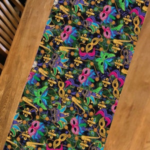 May include: A black table runner with a repeating pattern of colorful Mardi Gras masks, gold beads, and fleur-de-lis.