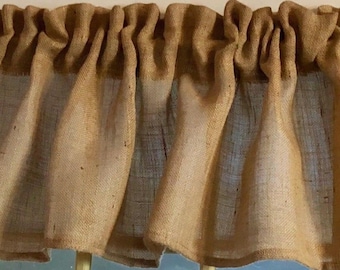 May include: A tan burlap valance with a gathered top edge. The fabric has a coarse texture and a natural, earthy appearance. The valance is designed to hang over a window, adding a rustic touch to home decor.