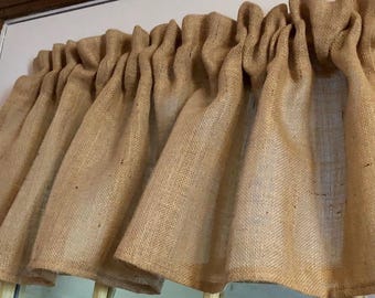 May include: A tan-colored window valance made of a coarse, woven burlap fabric. The valance features a gathered rod pocket top, creating vertical folds and pleats that drape downward across the width of the window frame in a uniform pattern.