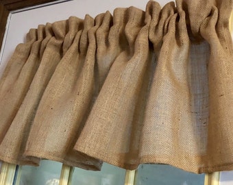 May include: A tan burlap window valance. The valance is gathered at the top, creating soft folds. The fabric has a coarse texture and a natural, rustic appearance. The valance is designed to be mounted above a window.