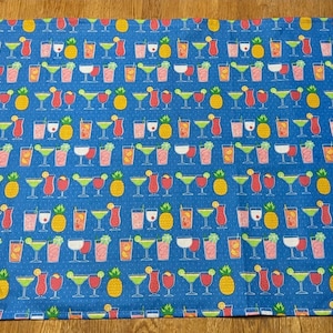 May include: A rectangular fabric kitchen towel featuring a repeating pattern of colorful cocktails and yellow pineapples on a bright blue background. The design includes various glasses filled with red, orange, and green drinks with garnishes.