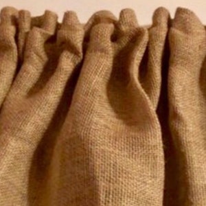 May include: A tan-colored curtain made of a coarse, woven fabric with a visible grid-like texture. The top edge of the curtain features a gathered rod pocket design, creating vertical folds that extend downward across the entire width.
