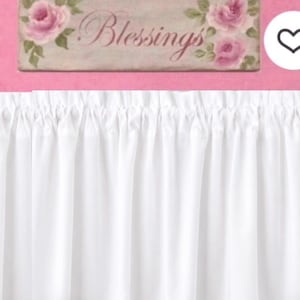 May include: A white curtain with a gathered top header. Above the curtain is a rectangular sign featuring a light beige background, pink rose illustrations at the corners, and the word Blessings written in a cursive, dark pink script font.
