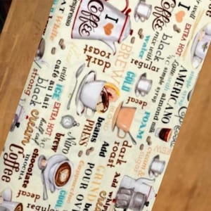 May include: A cream-colored fabric featuring a repeating pattern of coffee-themed illustrations and text. The design includes various coffee cups, mugs, and pots alongside words like I Love Coffee, Americano, Nitro brew, dark roast, and mocha in brown and blue.