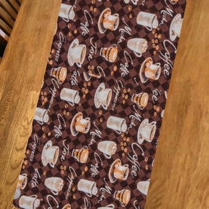 May include: A brown table runner with a repeating pattern of coffee cups and coffee beans. The cups are filled with different types of coffee, including cappuccino, latte, mocha, and espresso. The text "Coffee" is repeated throughout the pattern.