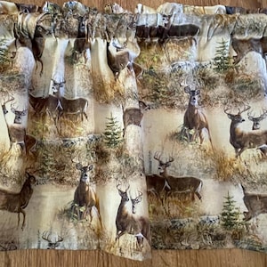 May include: A valance featuring a repeating pattern of deer in a naturalistic forest setting. The fabric has a beige background with brown and green details, creating a rustic, outdoorsy aesthetic. The valance is designed to be hung above a window.