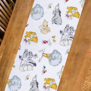 May include: White table runner with a repeating pattern of Winnie the Pooh and his friends, including Piglet, Eeyore, and Tigger. The characters are depicted in a cartoon style and are surrounded by flowers and trees.