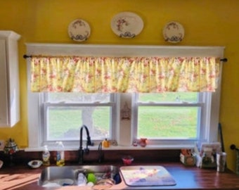 Può includere: Una finestra da cucina con una tenda gialla con un motivo floreale. La finestra ha due vetri e si affaccia su un prato verde. Un lavello in acciaio inox si trova sotto la finestra.
