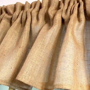 May include: A tan-colored window valance made of a coarse, woven burlap fabric. The valance features a gathered, ruffled top section that creates vertical folds and drapes downward, showcasing the natural, textured, and semi-sheer weave of the material.