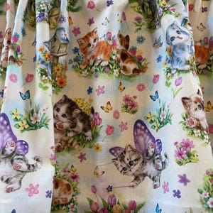 May include: Fabric with a whimsical print featuring kittens, butterflies, flowers, and birdhouses. The kittens are in various poses, some with fairy wings. The color palette includes pastels like pink, blue, and yellow.