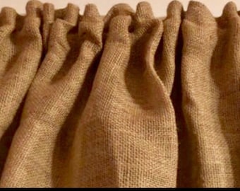 May include: Close-up of a tan burlap curtain valance. The fabric is woven with a visible texture and hangs in soft folds. The valance is mounted on a rod, creating a gathered effect.