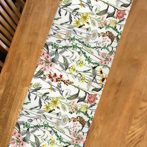 May include: A floral table runner with a white background and a pattern of pink, yellow, and red flowers, green vines, and gray leaves. The runner is placed on a wooden table.