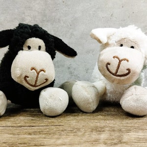 May include: Two plush sheep toys, one black and one white, sitting on a wooden surface. Both sheep have a brown embroidered smile and a white face.