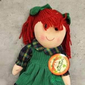 May include: A red-haired rag doll wearing a green plaid shirt and a green corduroy dress. The doll has a green shamrock on her dress and a tag that says "Roisin The cuddly Irish rag doll".