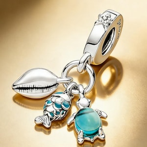 May include: A silver charm bracelet pendant featuring a seashell, a fish with blue accents, and a turtle with a blue gemstone shell. The pendant has a ring and a clasp with the word "Dream" engraved on it.