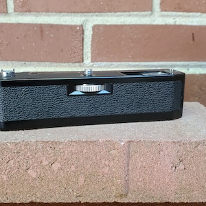 Puede incluir: Un dorso de cámara negro con una superficie texturizada y una perilla de rebobinado plateada.