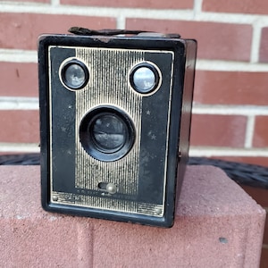 May include: A black vintage box camera with a lens and two viewfinders. The camera has a striped design and a gold trim.