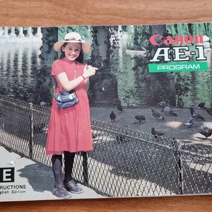 Op de afbeelding: Een vintage Canon AE-1 Program camera handleiding met een rood, wit en blauw omslag. De omslag toont een jong meisje in een rode jurk en een witte hoed die voor een hek staat met duiven op de achtergrond. De tekst "Canon AE-1 Program" is in wit op een groene achtergrond gedrukt.