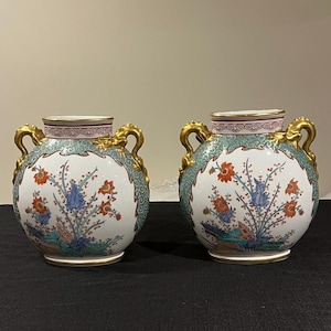 Puede incluir: Dos jarrones de porcelana con detalles dorados y diseños florales. Los jarrones tienen un fondo azul verdoso y blanco con motivos florales rojos, azules y verdes. Los jarrones tienen asas doradas y un borde dorado.
