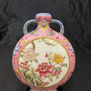 以下が含まれることがあります： ピンクの背景に2つのハンドルが付いた、丸くて平らな磁器製の花瓶。中央には、色鮮やかな牡丹と葉が描かれた白い円形のパネルがあります。首とベース部分は、複雑な花柄とスクロール模様で装飾されています。