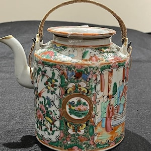 Puede incluir: Una tetera de porcelana vintage pintada a mano con un asa tejida. La tetera presenta un colorido diseño floral y figurativo con acentos de rosa, verde y dorado. El pico y la tapa son blancos.