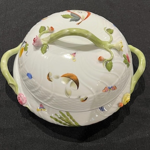 May include: White ceramic serving dish with a lid, adorned with colorful floral and mushroom designs. The lid features a green handle and is decorated with pink roses, butterflies, and other botanical elements. The dish has two green handles.
