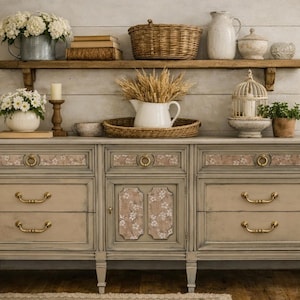 May include: A gray and beige painted wooden sideboard with floral accents and brass hardware. The top shelf holds a basket, books, and a metal bucket with flowers. A white pitcher and decorative items sit on the sideboard.