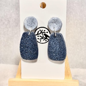 以下が含まれることがあります： 手作りのイヤリング。青い斑点模様のデザインです。各イヤリングは、小さく丸い上部と、より大きく長方形の下部が特徴です。イヤリングは、小さな木製のイーゼルに取り付けられた白いカードに表示されています。