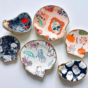 May include: A collection of six seashell-shaped decorative dishes. The dishes feature various Halloween-themed designs, including skulls, pumpkins, ghosts, and floral patterns. Each dish has a gold-colored rim and unique interior artwork.