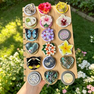 May include: A bamboo display board holding fifteen circular and heart-shaped decorative items. The collection features various designs including butterflies, hibiscus and plumeria flowers, an orchid, iridescent shell patterns, and textured stones.