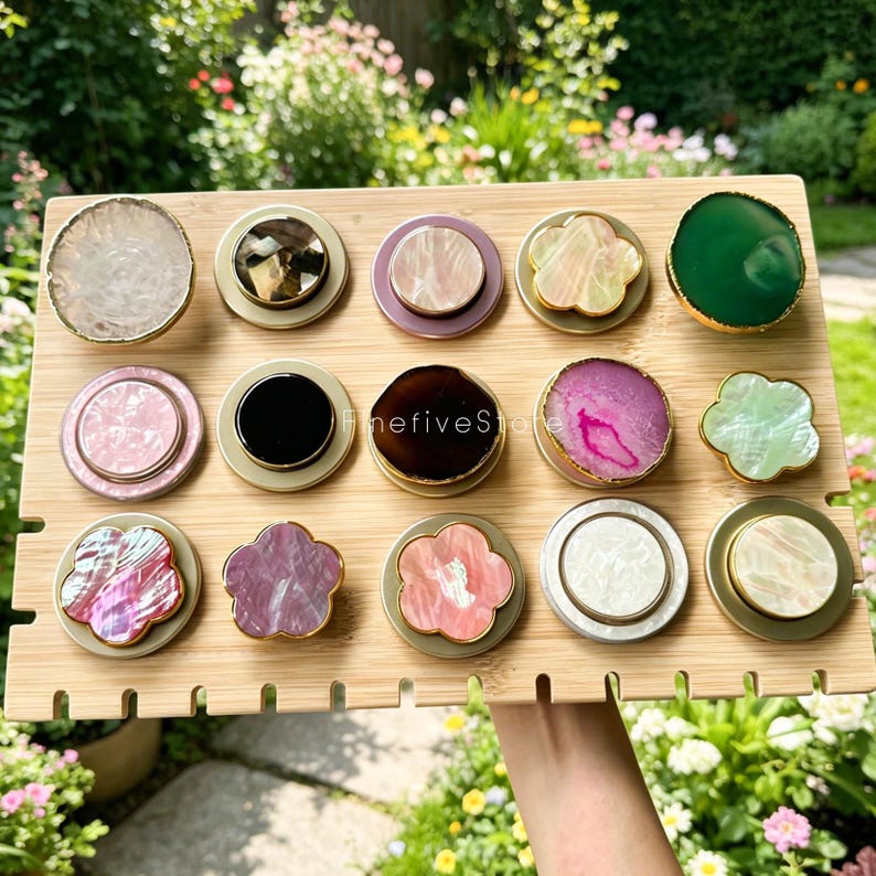 May include: A bamboo display board holds fifteen circular and flower-shaped phone grips with various stone, shell, and resin surfaces. The grips feature gold-toned edges and are arranged in three rows. The text finefiveStore is visible on one grip.