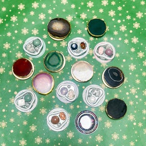 May include: A selection of decorative round objects with diverse colours and textures. Some have gold rims and feature natural stone or gemstone designs. The objects are arranged on a green background with white snowflake patterns.