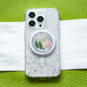Puede incluir: Funda transparente para teléfono con diseño floral y purpurina plateada. La funda tiene un agarre circular blanco con flores secas y vegetación. La funda del teléfono tiene una parte trasera transparente con diseño de purpurina plateada y detalles de estrellas.