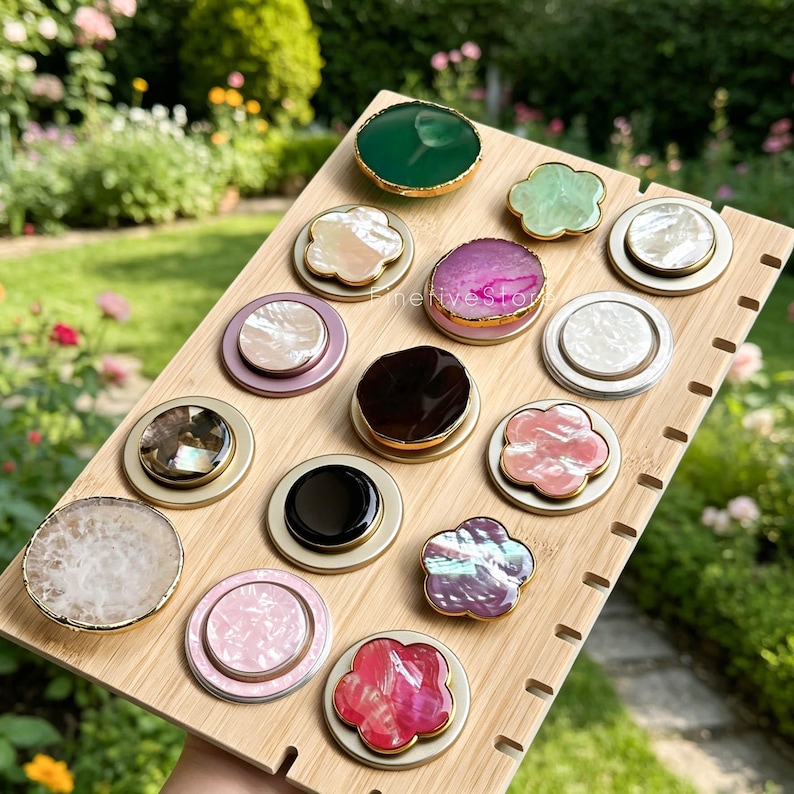 May include: A collection of circular and flower-shaped phone grips arranged on a light wood display board. The grips feature various finishes, including polished stone, iridescent shell, solid black, and marbled patterns with gold-toned metal edges.