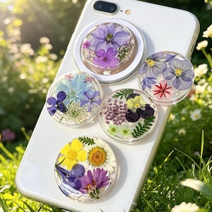 May include: Five circular phone grips attached to a white smartphone, each featuring a clear resin top embedded with various pressed flowers including purple, blue, yellow, and white petals, along with small green fern leaves and delicate foliage.
