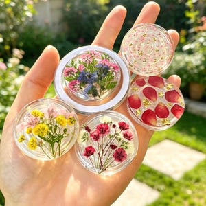 May include: Five circular clear resin accessories featuring embedded dried flowers and gold leaf flakes. One piece contains pink and blue flowers, another yellow and pink, one red and white, one red petals, and one small pink flowers with gold.