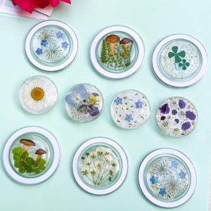 May include: A collection of clear, round resin coasters with white rims, each containing different dried flowers, mushrooms, and a four-leaf clover. The coasters feature blue, white, yellow, and purple floral arrangements, and gold flecks.