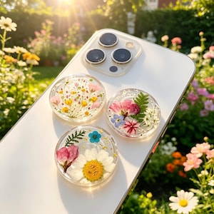 May include: Three circular phone grips attached to the back of a white smartphone. Each grip is made of clear resin containing pressed dried flowers, including small yellow, pink, and white blossoms, green fern leaves, and tiny gold flakes.
