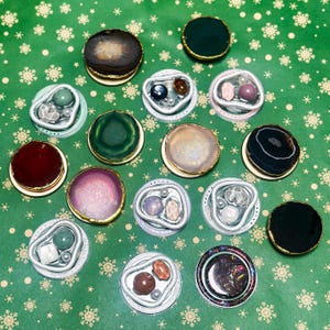May include: A selection of gemstone coasters with gold-coloured rims. The coasters display diverse colours and patterns, including green, purple, brown, and black. Some coasters have embedded stones and pearls. The background is green wrapping paper with white snowflake patterns.