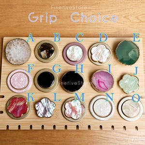 May include: A collection of fifteen circular and flower-shaped phone grips labeled A through O. The grips feature various materials including white, pink, black, and green stone, shell, and resin inlays with gold or silver-toned metal edges.