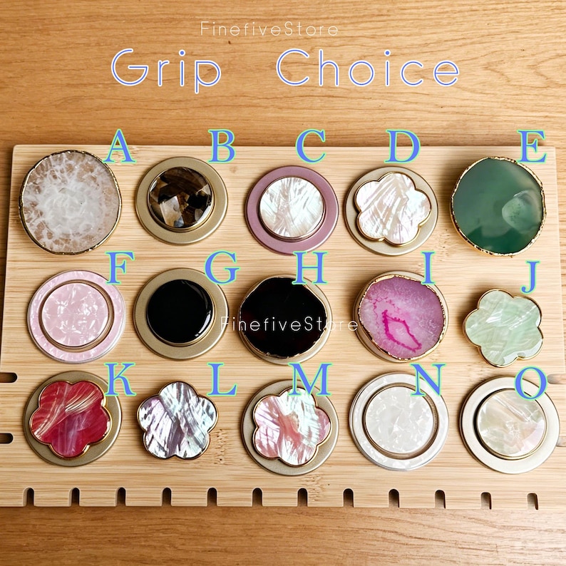 May include: A collection of fifteen circular and flower-shaped phone grips labeled A through O. The grips feature various materials including white, pink, black, and green stone, shell, and resin inlays with gold or silver-toned metal edges.