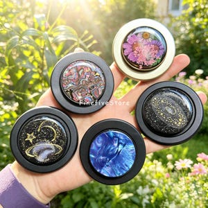 Può includere: Cinque supporti circolari per telefono con design distinti: uno con fiori pressati viola e rosa in una cornice dorata, uno con conchiglia di abalone iridescente, uno con vortice galattico blu, uno con motivo a stelle e uno con un pianeta dorato.