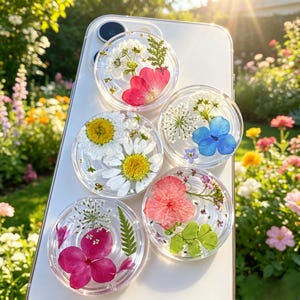 May include: A white smartphone case featuring four circular clear resin attachments, each embedded with various pressed flowers including white daisies, pink hydrangea petals, blue blossoms, and green fern leaves arranged in distinct patterns.