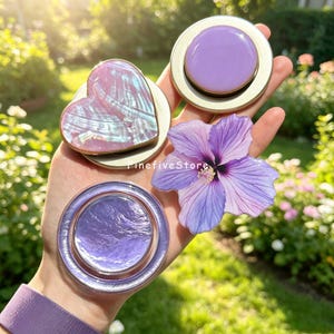 May include: A collection of four circular and heart-shaped phone grips featuring a variety of designs, including a pearlescent pink heart, a solid lavender circle, a textured purple metallic circle, and a light purple hibiscus flower graphic.