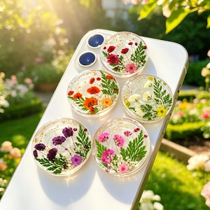 May include: Five circular phone grips attached to a white smartphone, each featuring clear resin embedded with pressed flowers and gold flakes. The designs include red, orange, white, purple, and pink blossoms paired with green fern leaves.