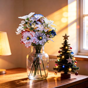 May include: A clear glass vase filled with colorful, decorative flowers. Next to the vase is a small Christmas tree decorated with lights and ornaments. The scene is set on a wooden surface, with a lamp and window in the background.