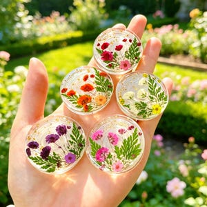 以下が含まれることがあります： 透明な樹脂製の円形スマホグリップ5個セット。押し花と緑のシダの葉が封入されています。各ピースには、赤、ピンク、紫、白、黄色の花々がそれぞれ異なる配置でデザインされており、金箔のような小さなフレークが散りばめられています。