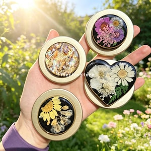 May include: Four circular and heart-shaped gold-toned metal phone grips featuring decorative inserts. One displays iridescent abalone shell, while three contain pressed flowers in pink, purple, white, and yellow on black backgrounds. Text reads FivefiveStore.
