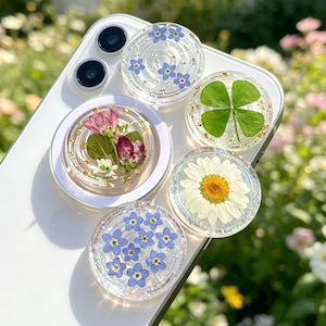 Pode incluir: Cinco suportes circulares para telemóvel com topos em resina transparente fixados num smartphone branco. Os suportes apresentam flores prensadas incrustadas, incluindo miosótis azuis, um trevo de quatro folhas, uma margarida e pétalas de rosa com flocos dourados.