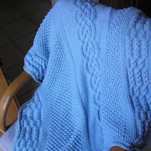 May include: A light blue knitted blanket with intricate cable and textured stitch patterns. The blanket is draped over a wooden chair, showcasing the detailed craftsmanship. The cable knit design runs along the edges and center of the blanket.